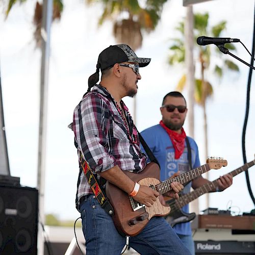 Two musicians are playing electric guitars on an outdoor stage. Palm trees are visible in the background, and they both wear casual clothes and sunglasses.