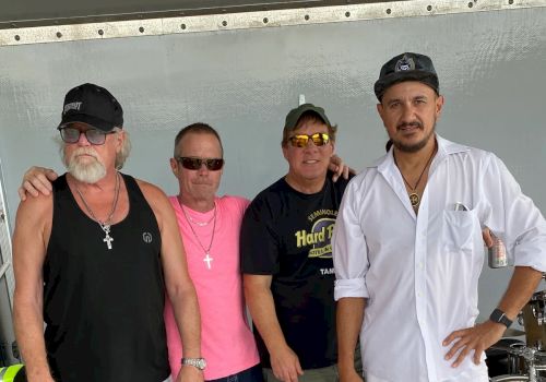 Four men pose by stage gear, including a chef in a white uniform and three in casual hats and sunglasses, smiling.