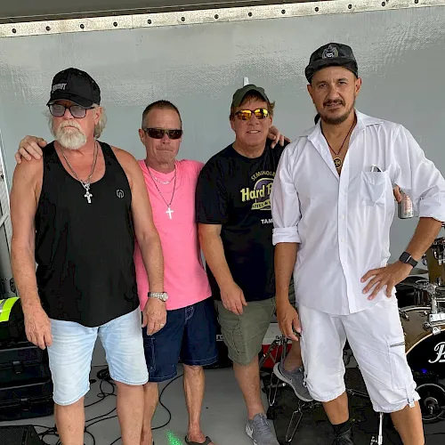 Four men pose by stage gear, including a chef in a white uniform and three in casual hats and sunglasses, smiling.