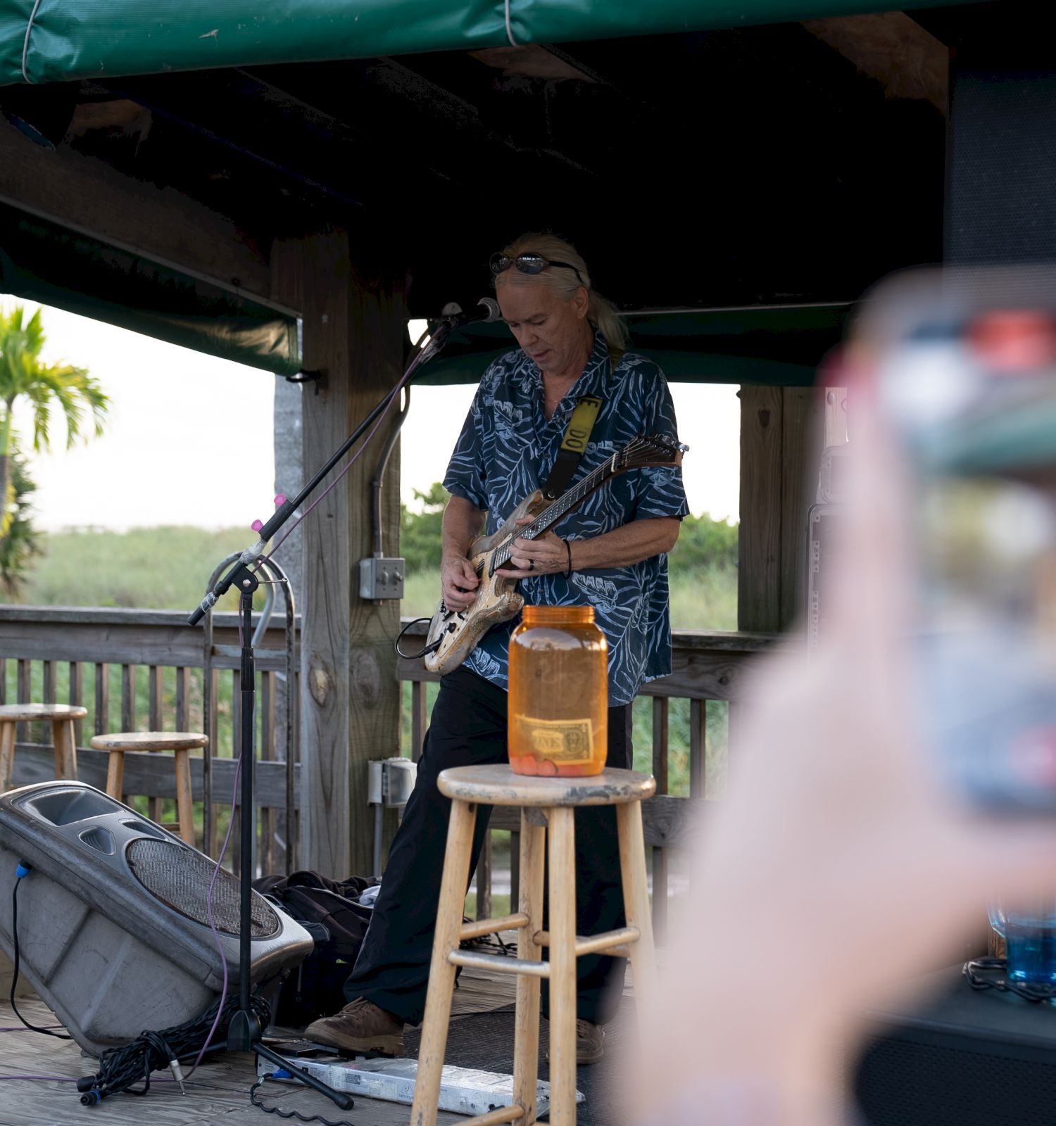 A musician is playing a guitar in an outdoor wooden structure, while someone takes a photo with a smartphone in the foreground.