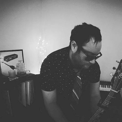 A man wearing sunglasses and a tie plays a guitar in a room with a keyboard and framed artwork in the background.