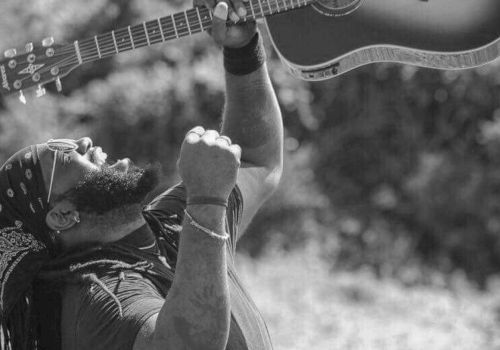 A person is outdoors, enthusiastically lifting an acoustic guitar overhead with both hands, while their mouth is open, possibly shouting or singing.