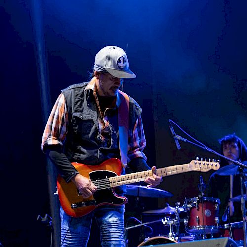 A man is playing an electric guitar on stage, wearing a hat and casual clothes, with a drummer in the background. The lighting is blue.