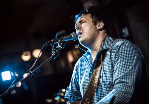 A person is passionately singing into a microphone while playing a guitar, under stage lighting in a performance setting, wearing a checkered shirt.