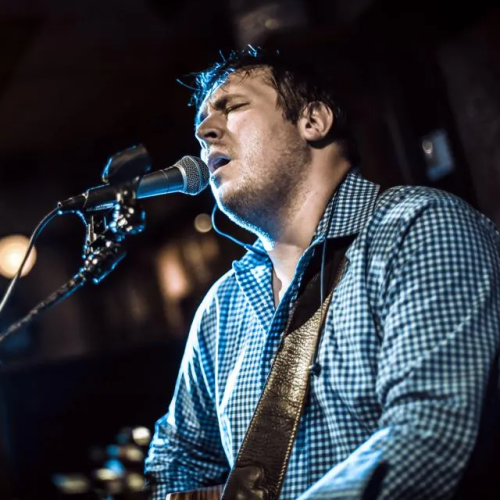 A person is passionately singing into a microphone while playing a guitar, under stage lighting in a performance setting, wearing a checkered shirt.