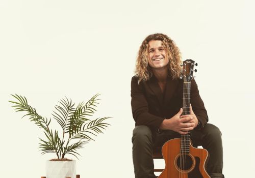 A man with curly hair sits on a chair, holding an acoustic guitar, with a potted plant on a stand to the left in a minimalist setting.