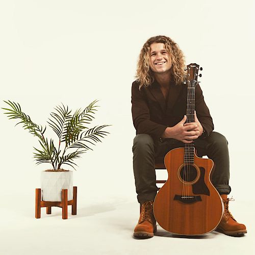 A man with curly hair sits on a chair, holding an acoustic guitar, with a potted plant on a stand to the left in a minimalist setting.