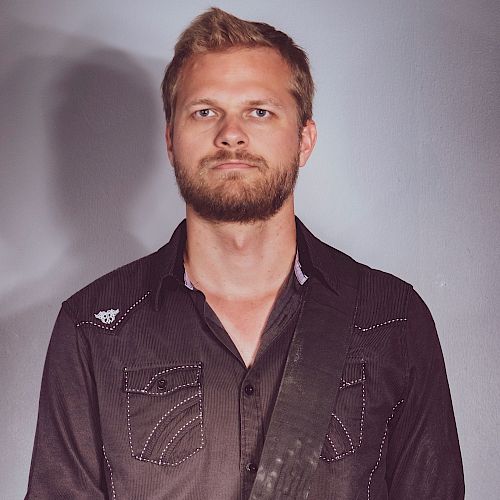 A bearded man with short hair stands against a plain background, wearing a dark button-up shirt with a patterned design and a strap across his chest.