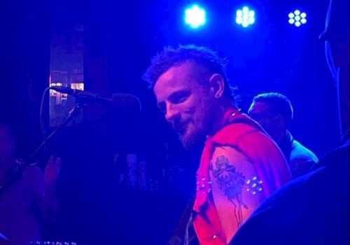 A musician with a beard and red vest playing an electric guitar on stage with blue lighting in the background, facing toward another musician.