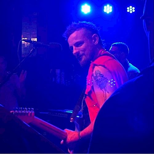 A musician with a beard and red vest playing an electric guitar on stage with blue lighting in the background, facing toward another musician.