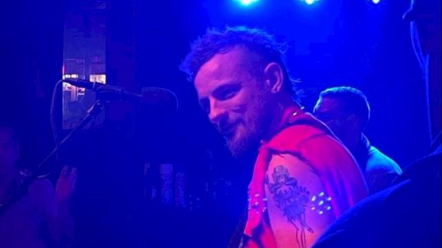 A musician with a beard and red vest playing an electric guitar on stage with blue lighting in the background, facing toward another musician.
