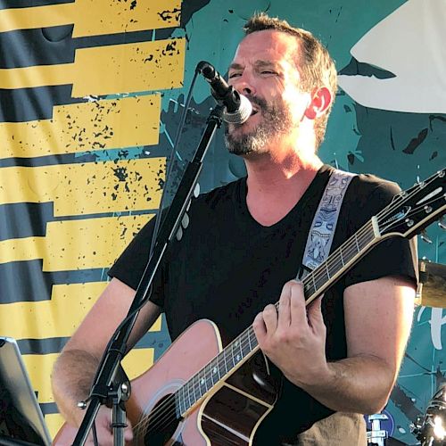 A man is singing into a microphone while playing an acoustic guitar on stage, with a backdrop featuring a yellow and blue design.