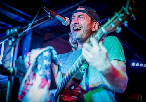 A musician is passionately singing and playing guitar on stage, wearing a cap, with a vibrant spotlight and an American flag detail.
