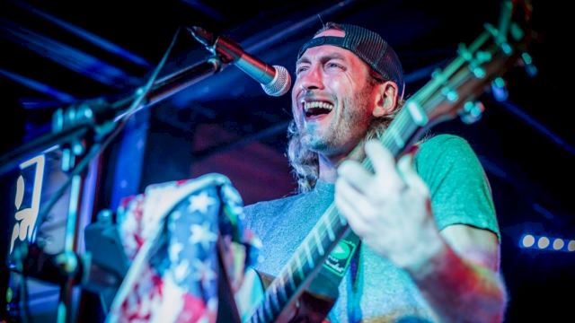 A musician is passionately singing and playing guitar on stage, wearing a cap, with a vibrant spotlight and an American flag detail.
