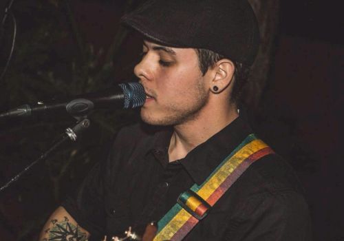 A person is singing into a microphone while playing an acoustic guitar, wearing a cap and a dark shirt with a multi-colored strap.