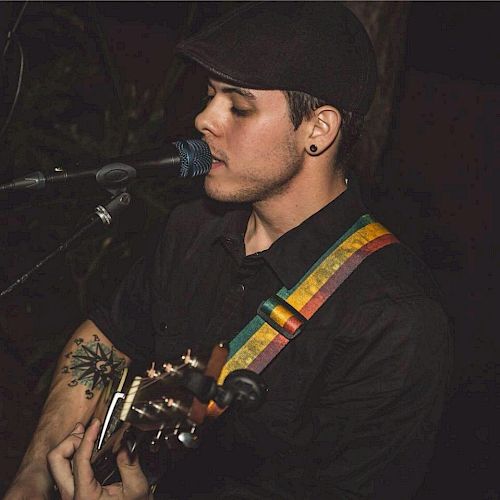 A person is singing into a microphone while playing an acoustic guitar, wearing a cap and a dark shirt with a multi-colored strap.