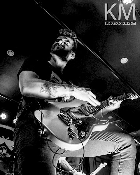 A man is passionately playing an electric guitar on stage, captured in black and white. The photo is credited to KM Photography.