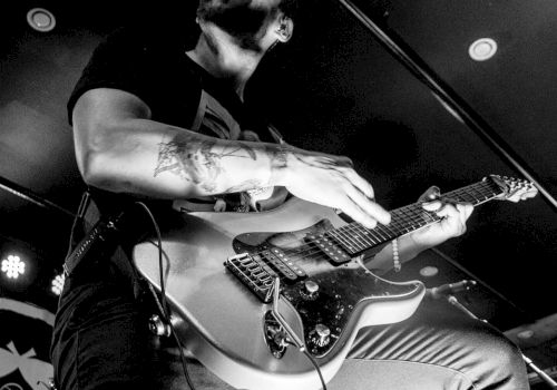 A man is passionately playing an electric guitar on stage, captured in black and white. The photo is credited to KM Photography.