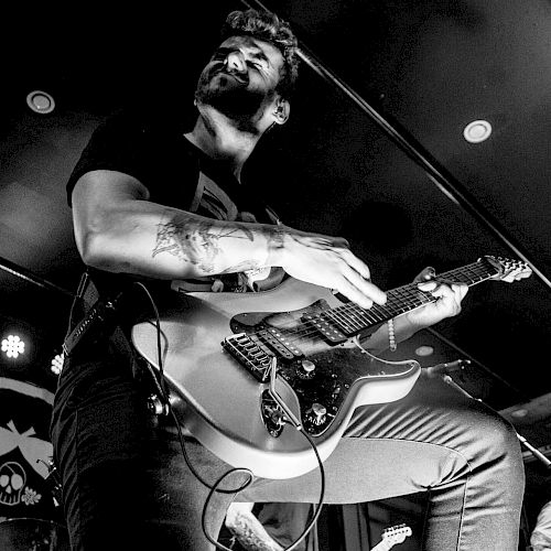 A man is passionately playing an electric guitar on stage, captured in black and white. The photo is credited to KM Photography.