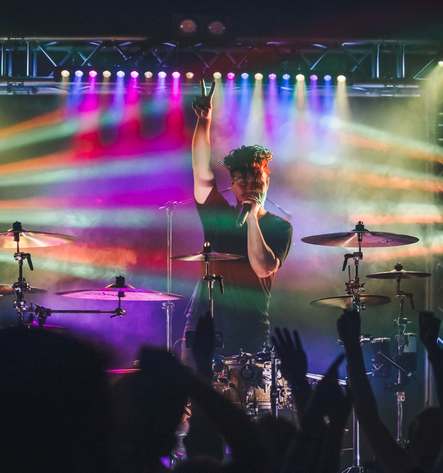 A drummer performs on stage with colorful lights and a cheering crowd in the foreground.