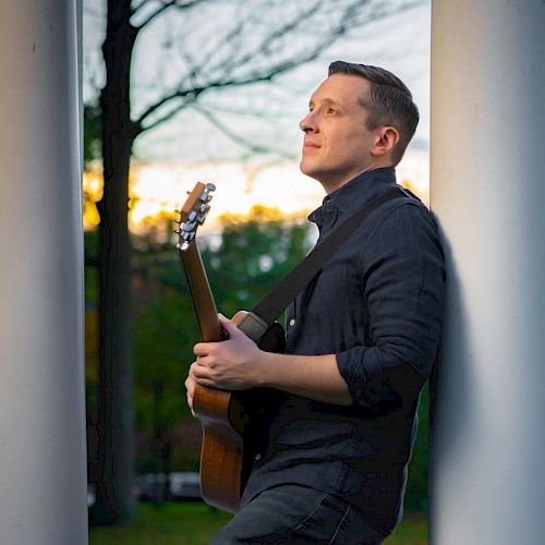 A person is leaning against a white pillar, holding a guitar, in an outdoor setting with trees and a sunset in the background.