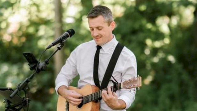 A person in formal attire is playing an acoustic guitar outdoors, with a microphone and stand nearby, surrounded by greenery.