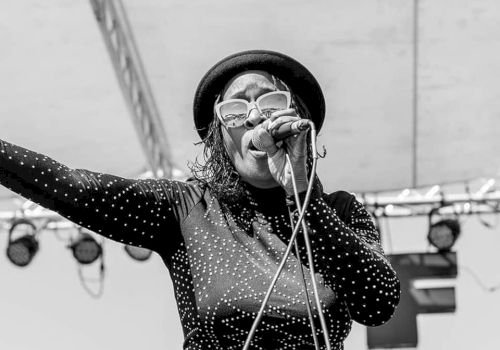 A  lady wearing a hat and glasses is singing into a microphone on a stage. The photo credits 