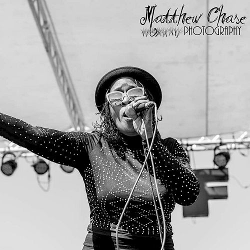 A  lady wearing a hat and glasses is singing into a microphone on a stage. The photo credits 