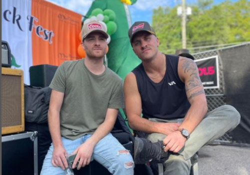 Two men are sitting together at an outdoor event, one holding a bottle. There is an inflatable dinosaur in the background.