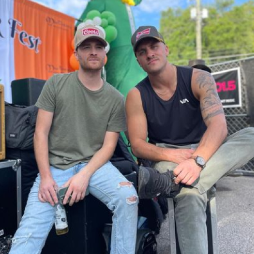 Two men are sitting together at an outdoor event, one holding a bottle. There is an inflatable dinosaur in the background.