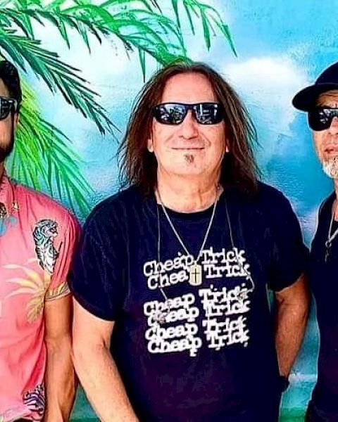 Three men wearing sunglasses stand in front of a tropical mural with a palm tree. One wears a floral shirt, and another wears a 