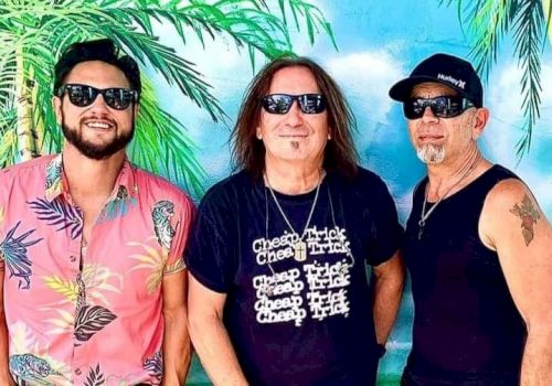 Three men wearing sunglasses stand in front of a tropical mural with a palm tree. One wears a floral shirt, and another wears a 