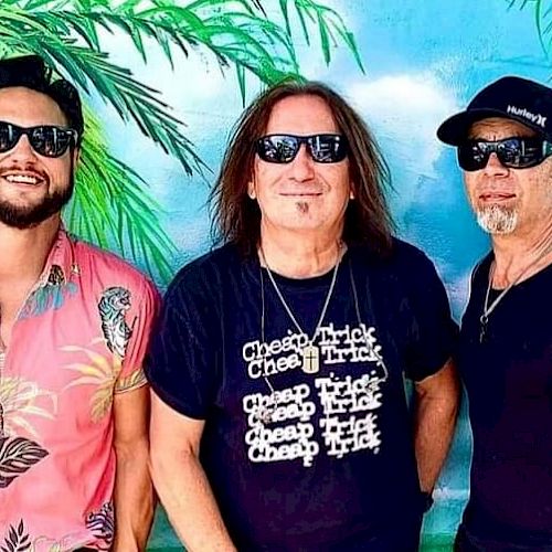 Three men wearing sunglasses stand in front of a tropical mural with a palm tree. One wears a floral shirt, and another wears a 