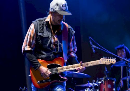 A guitarist wearing a hat performs on stage under blue lights, with a drummer partially visible in the background.