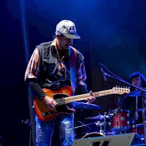 A guitarist wearing a hat performs on stage under blue lights, with a drummer partially visible in the background.