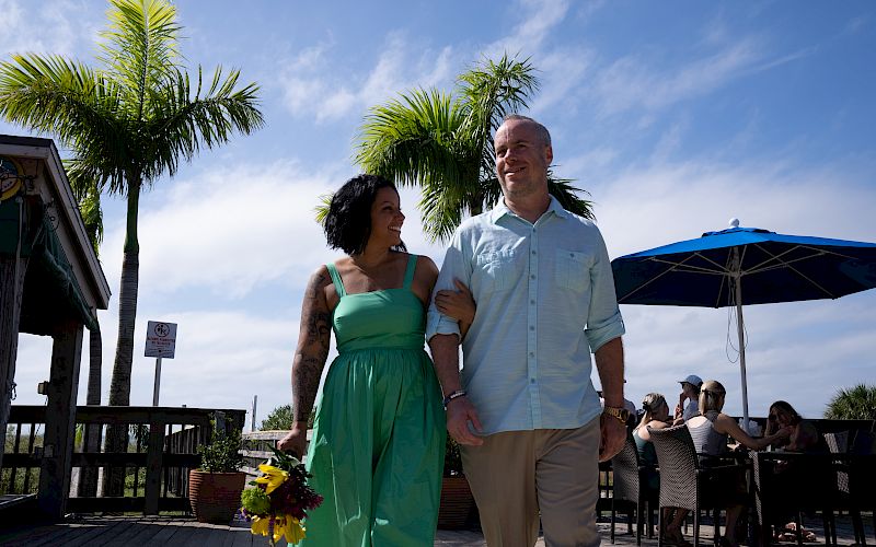 A couple is walking arm-in-arm near outdoor seating by palm trees. One person is holding a bouquet of flowers under a bright, sunny sky.