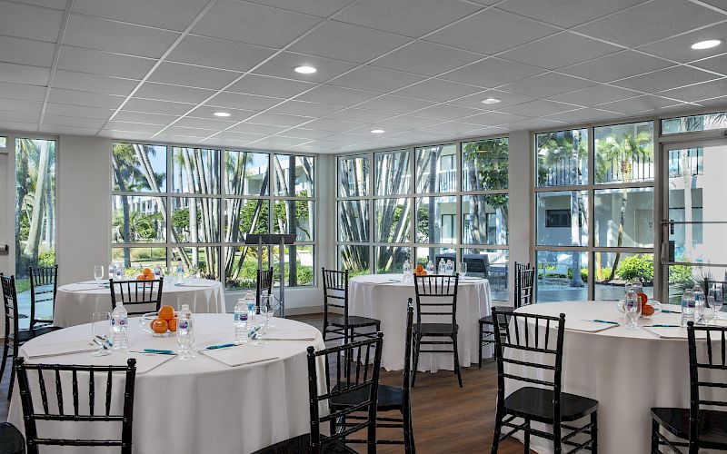 A conference room with round tables covered in white tablecloths, black chairs, and windows with a view of a garden outside. The sentence ends.