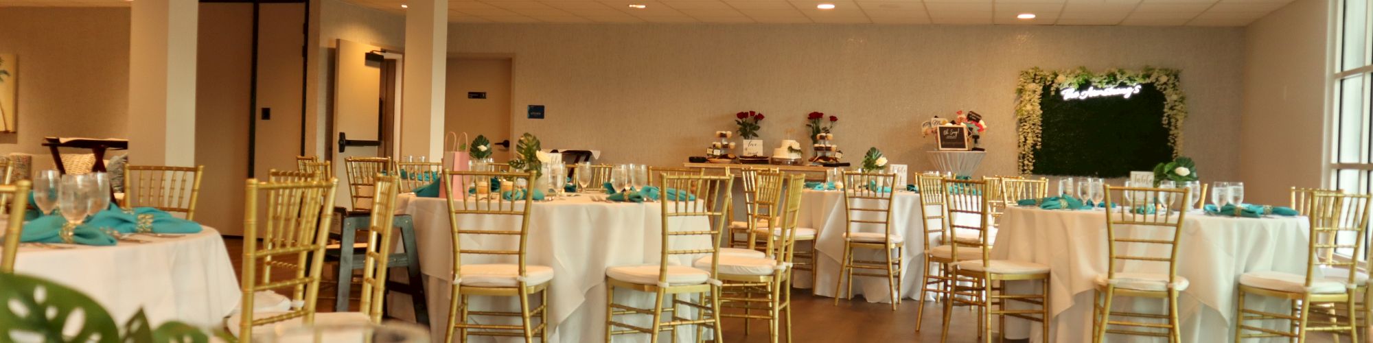 A decorated indoor event space with white tablecloths, gold chairs, and green accents. Tables have place settings and candles, with a backdrop.