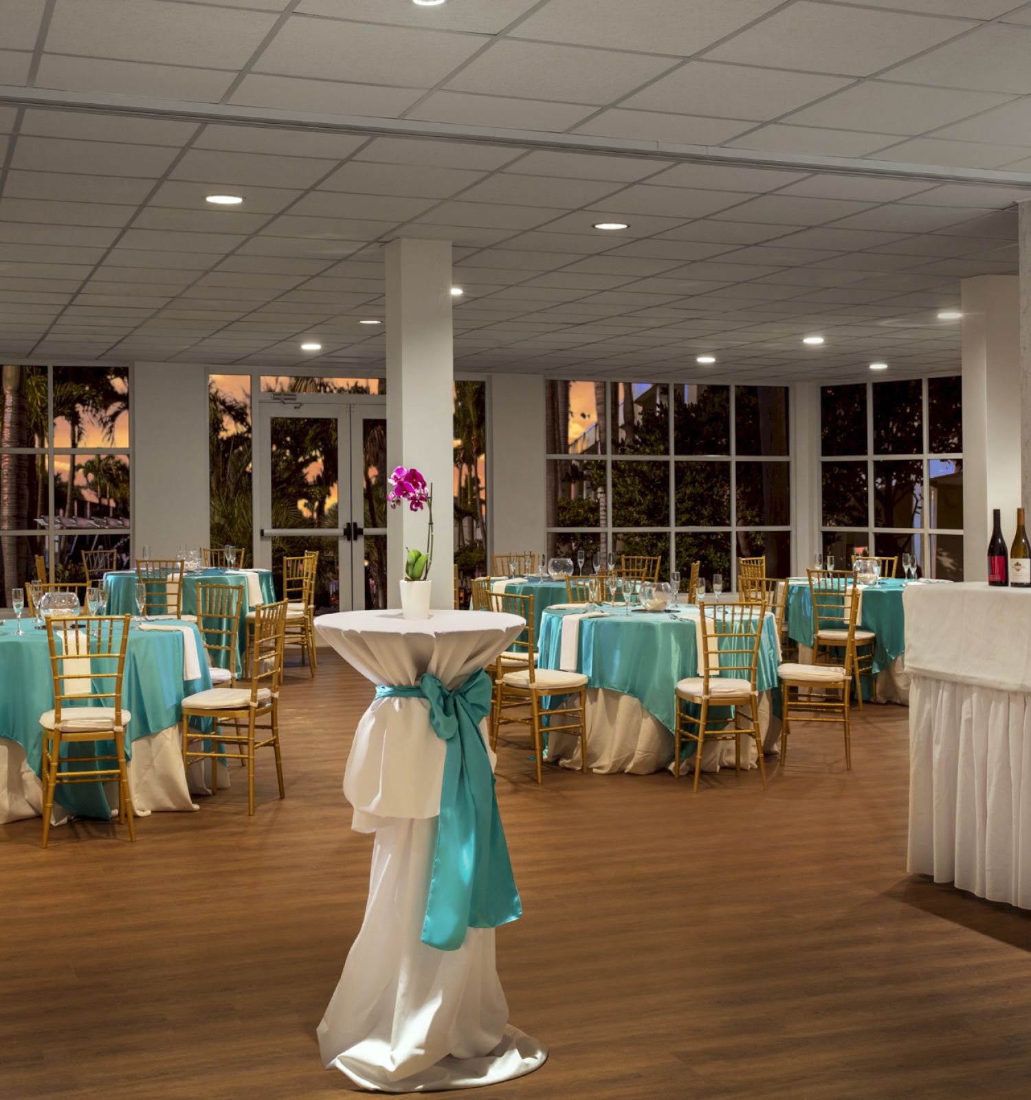 The image shows a decorated banquet hall with round tables covered with turquoise tablecloths, adorned chairs, and high tables with white and turquoise bows.