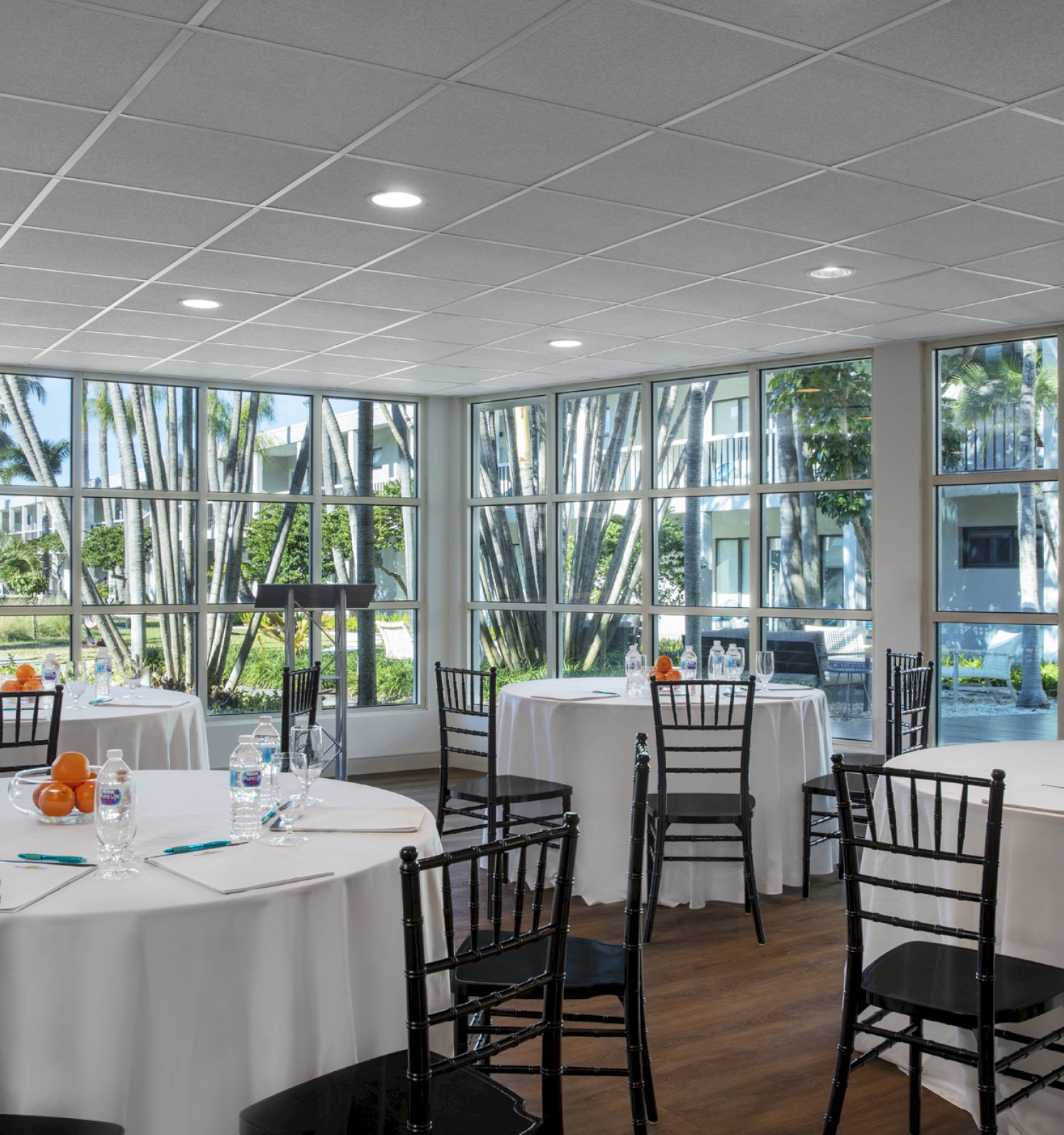 The image shows a bright room set up for an event with round tables, white tablecloths, black chairs, and fruits as centerpieces, surrounded by large windows.