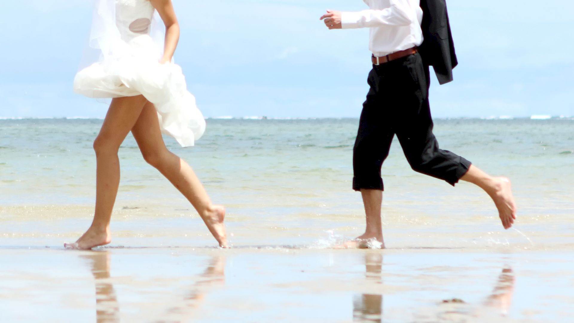 A bride and groom are walking barefoot on a beach. The bride is wearing her wedding dress, and the groom is in a shirt with his jacket slung over his shoulder.