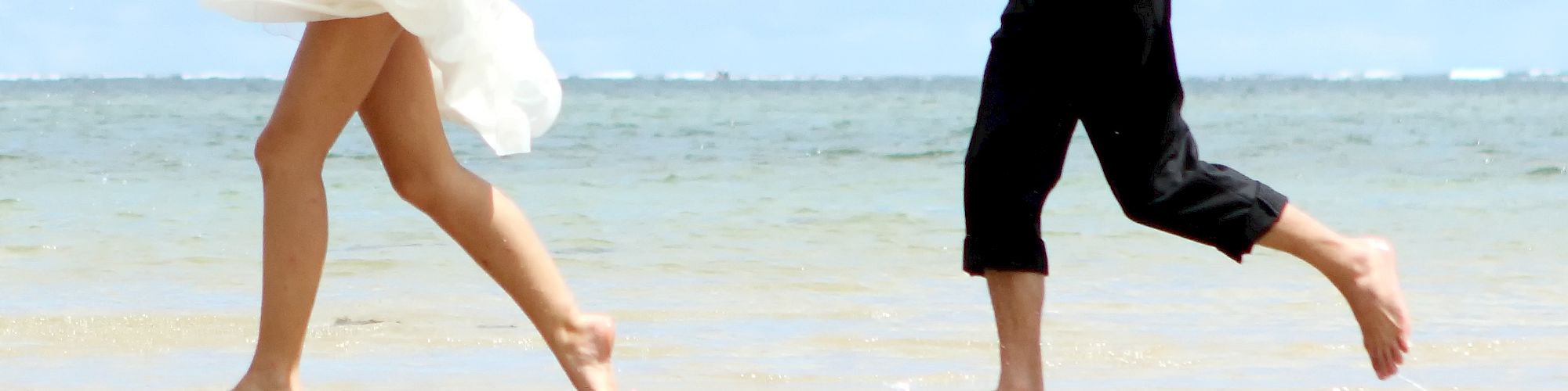 A bride and groom are walking barefoot on a beach. The bride is wearing her wedding dress, and the groom is in a shirt with his jacket slung over his shoulder.