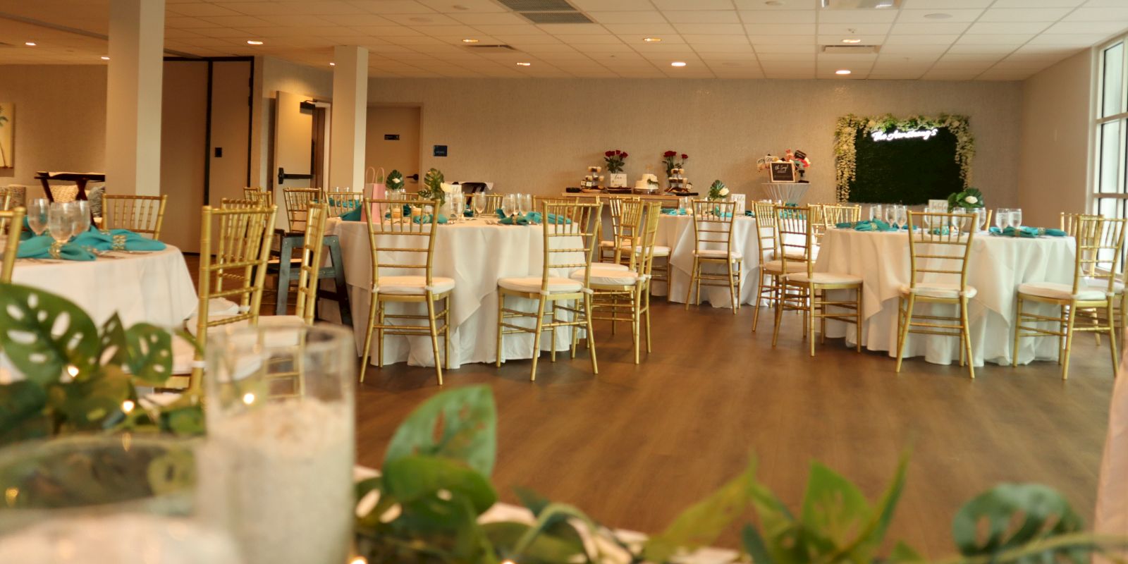 The image shows a decorated event space with white tablecloths, gold chairs, and green accents, possibly set up for a wedding or banquet.