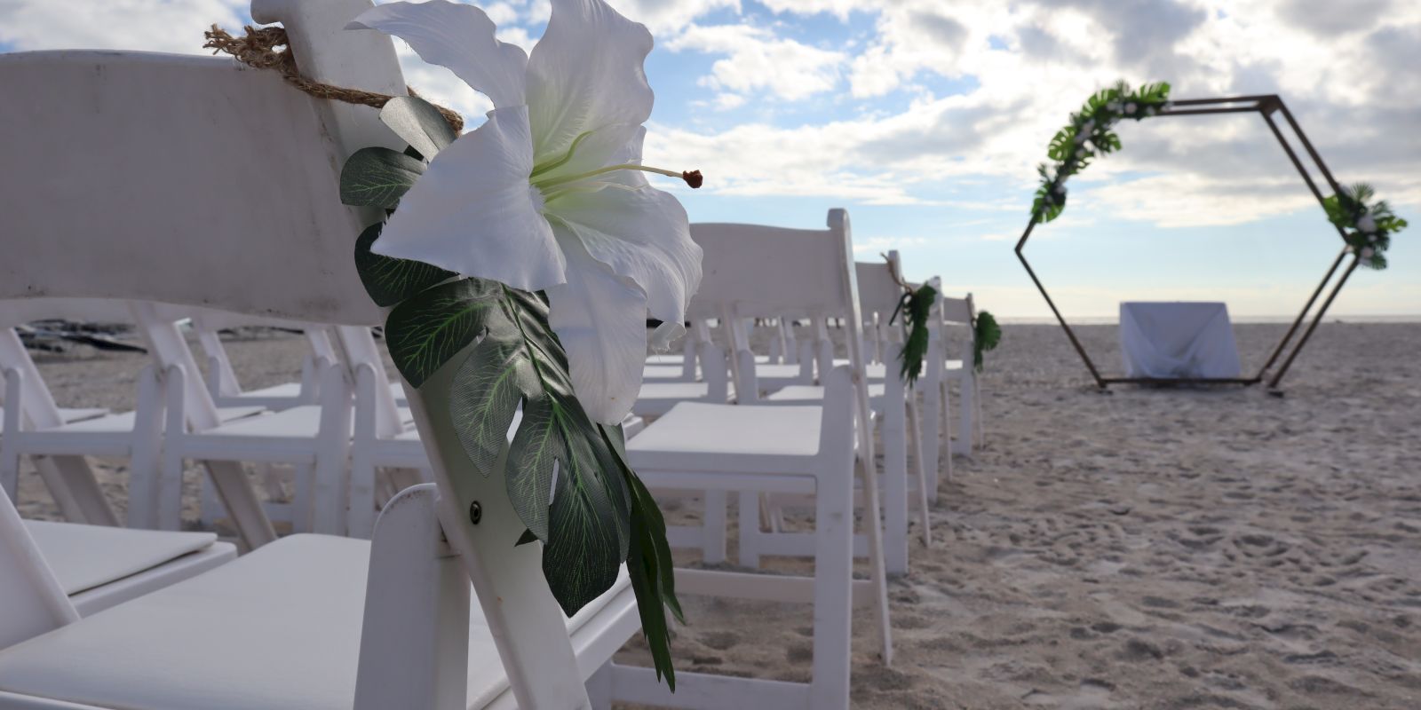 This image shows a beach wedding setup with white chairs, each adorned with white flowers and leaves, leading to a hexagonal arch decorated with greenery.