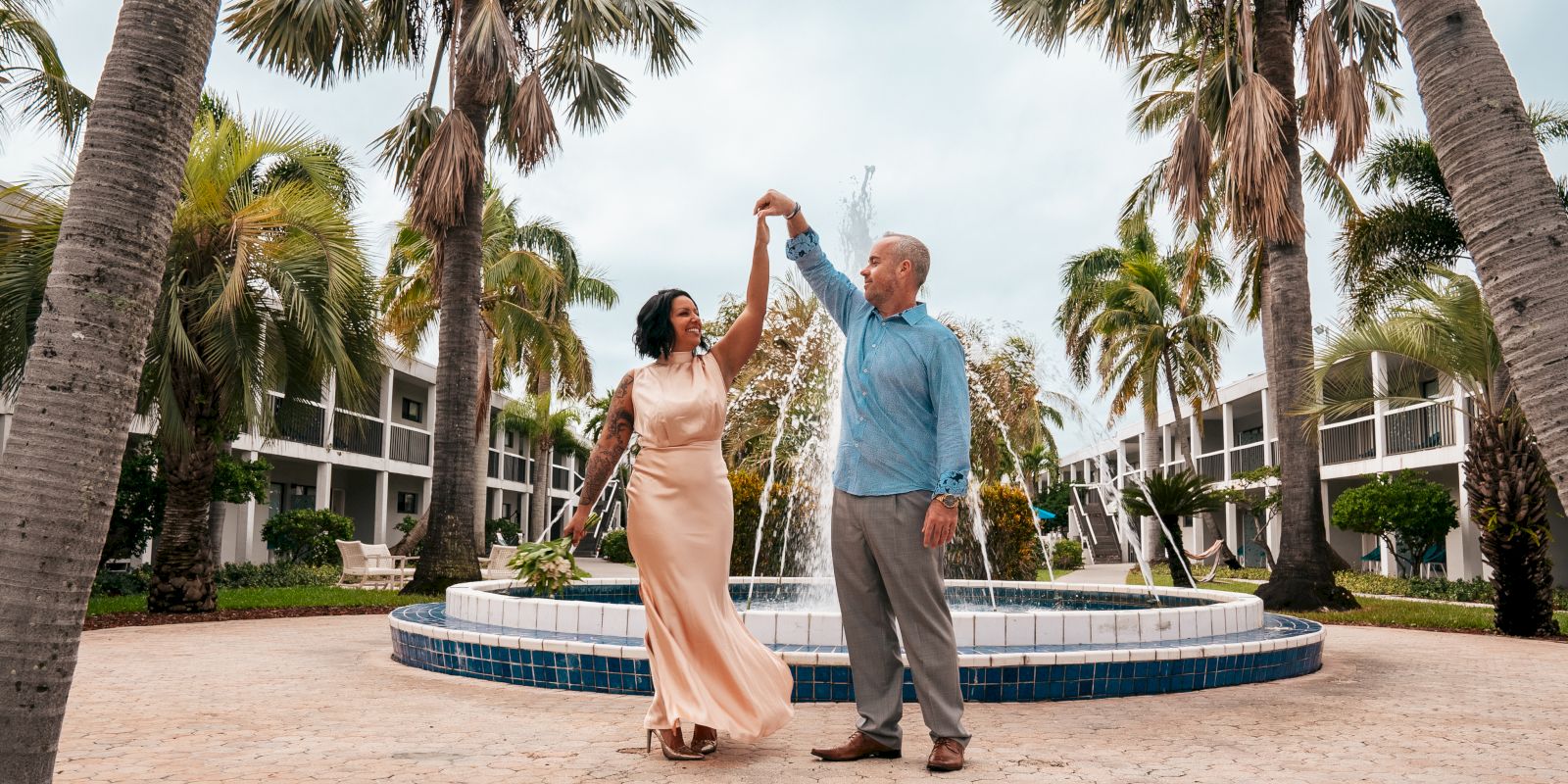 A couple dances near a fountain surrounded by palm trees and buildings on a paved area, creating a serene and romantic atmosphere.