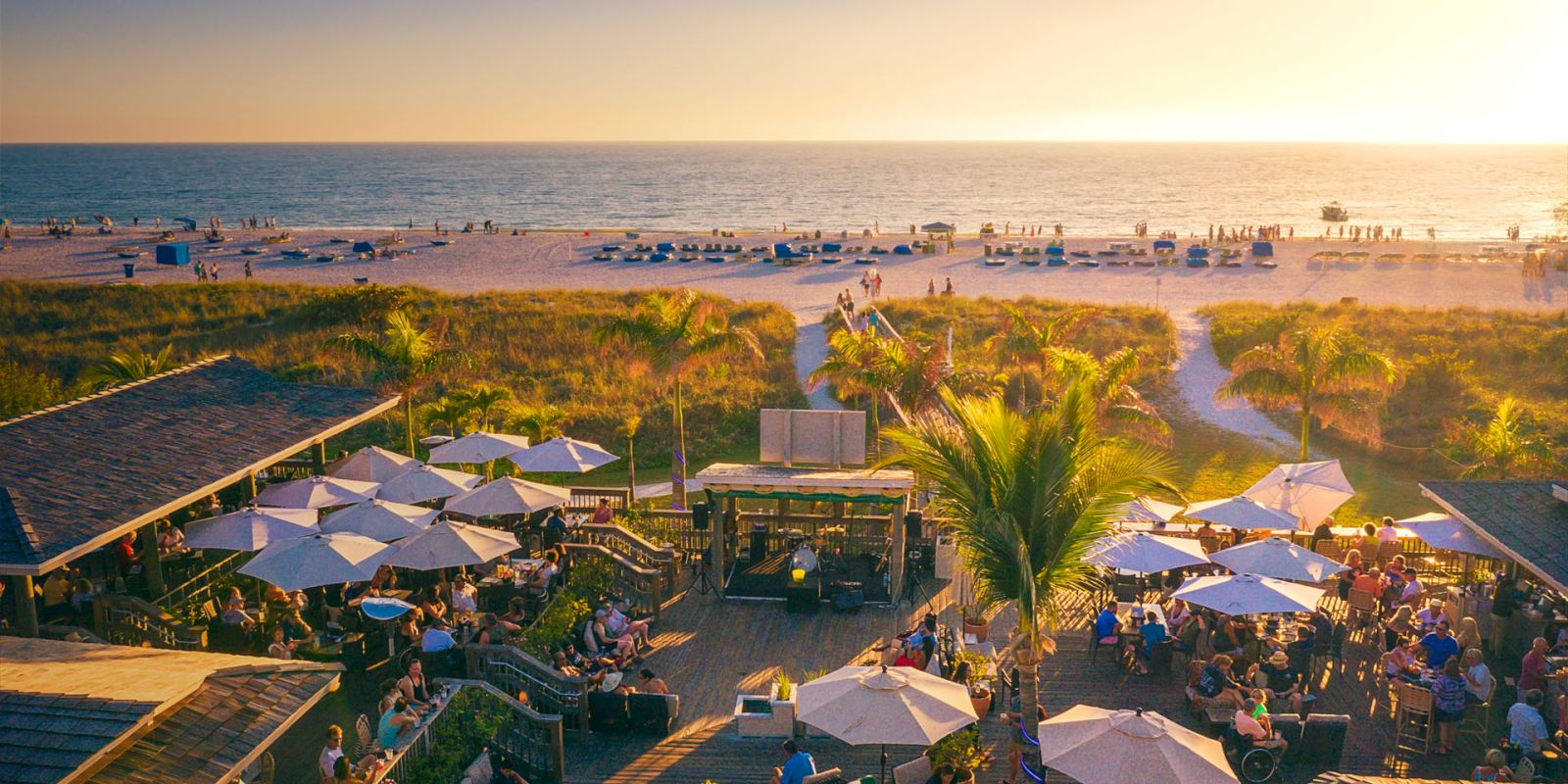 The image shows a beachside restaurant or bar with outdoor seating, filled with people enjoying the sunset, surrounded by palm trees and beach views.