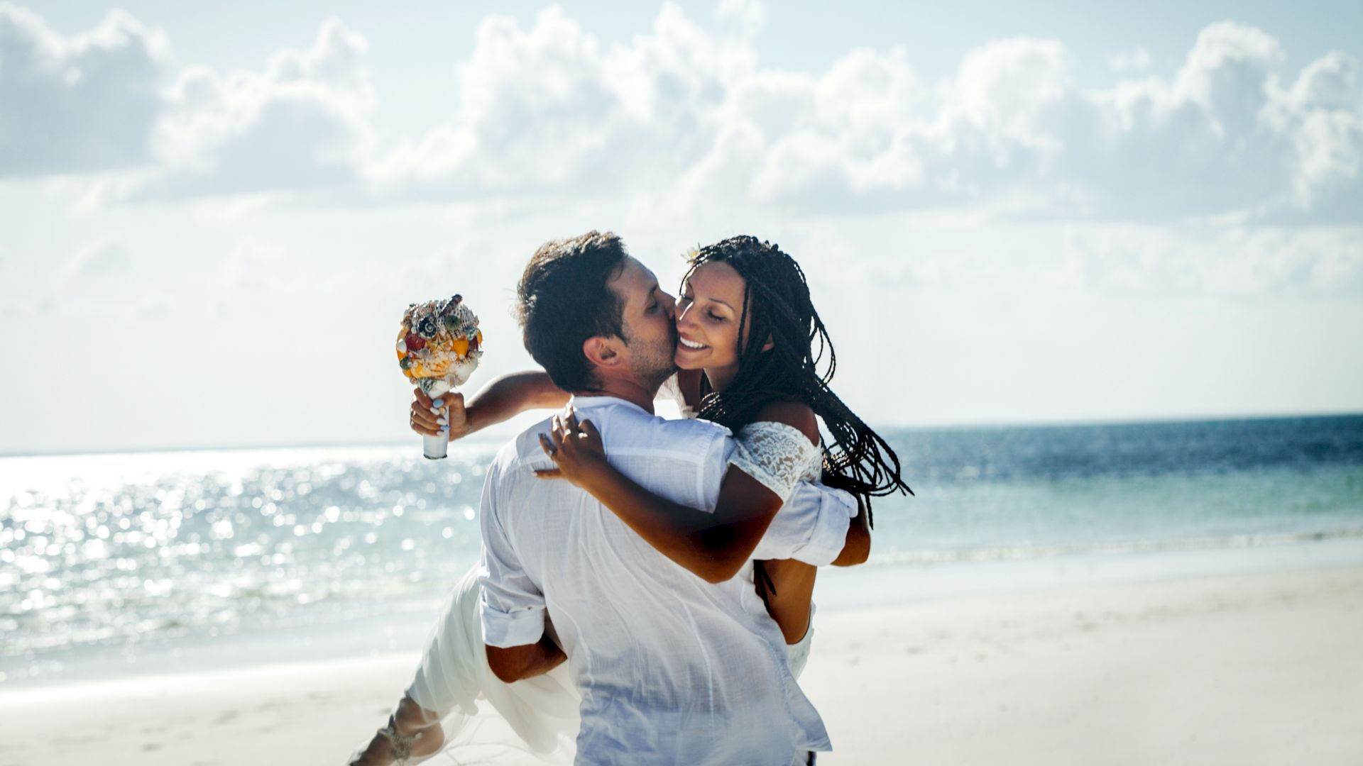 A person is carrying another person and holding a bouquet on a beach. Both people appear to be joyful under a partly cloudy sky, near the ocean.