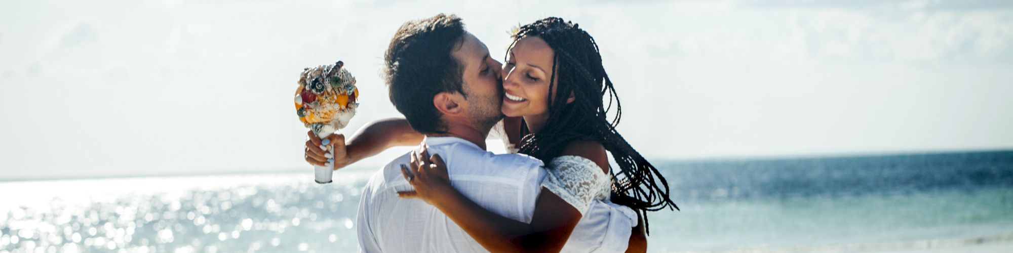 A person is carrying another person and holding a bouquet on a beach. Both people appear to be joyful under a partly cloudy sky, near the ocean.