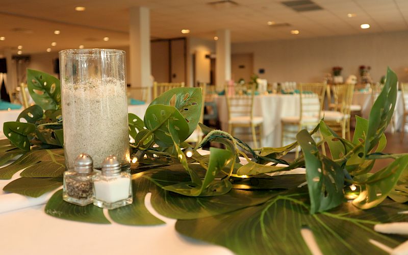 The image shows a decorated table with green leaves, a large candle, salt and pepper shakers, and a banquet hall with chairs in the background.