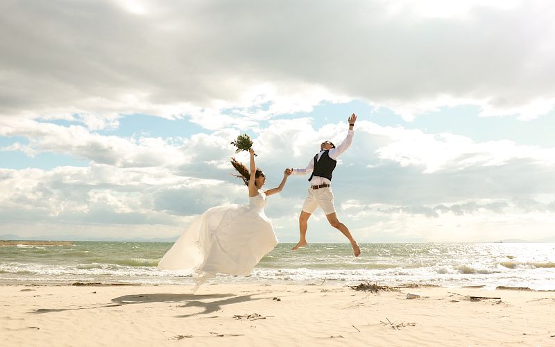 A bride and groom jump joyfully on a sandy beach with the ocean and cloudy sky in the background, holding hands and celebrating.